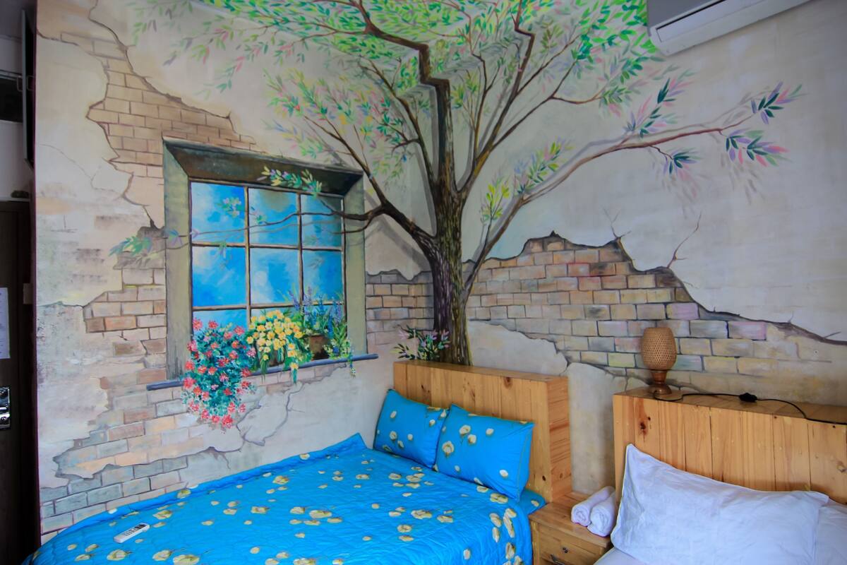 A bedroom with a mural of a tree, a window with flowers, a blue bedspread, and a wooden headboard.