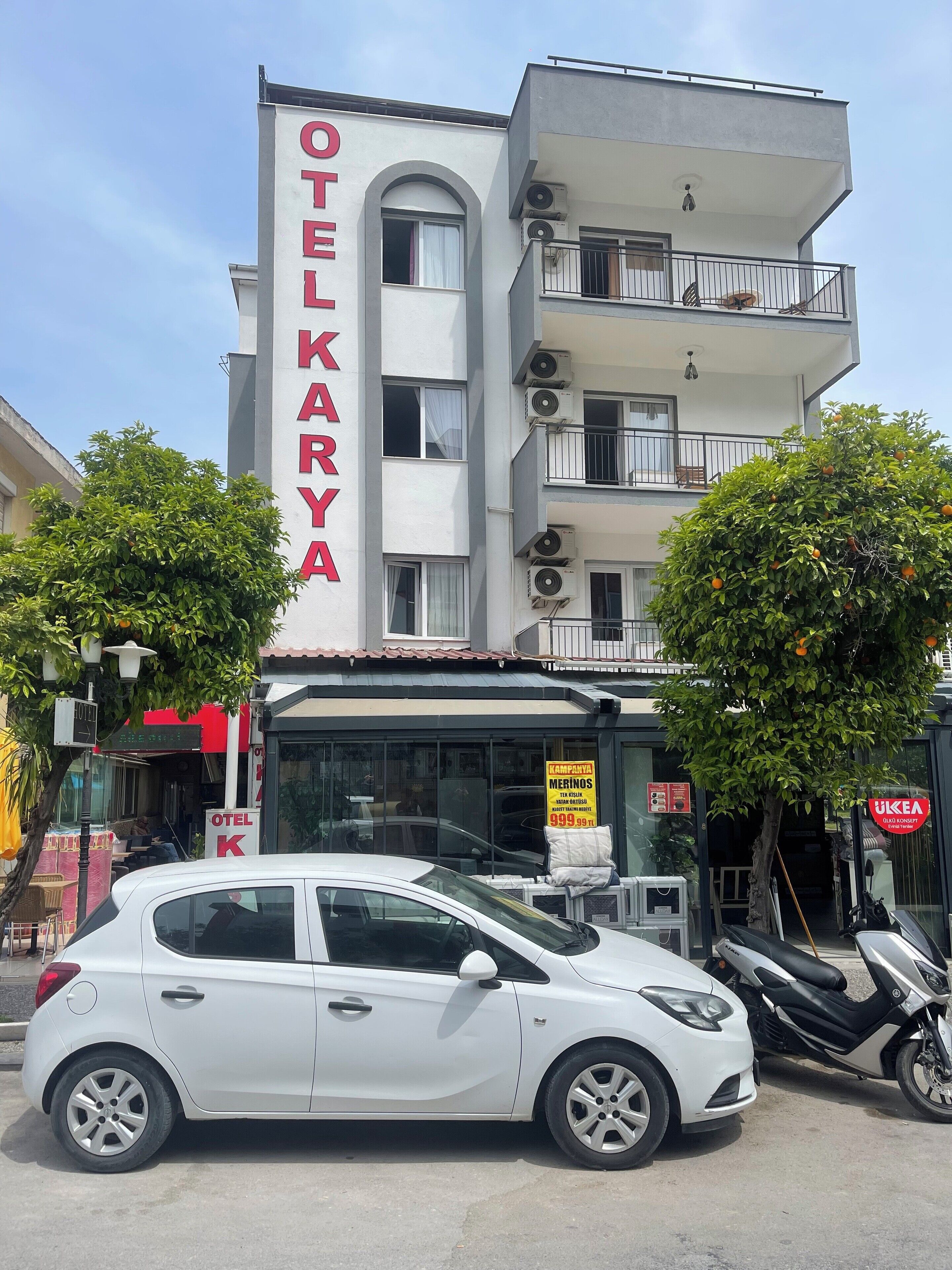A hotel named "OTEL KARYA" with a modern facade, multiple balconies, and parked cars.