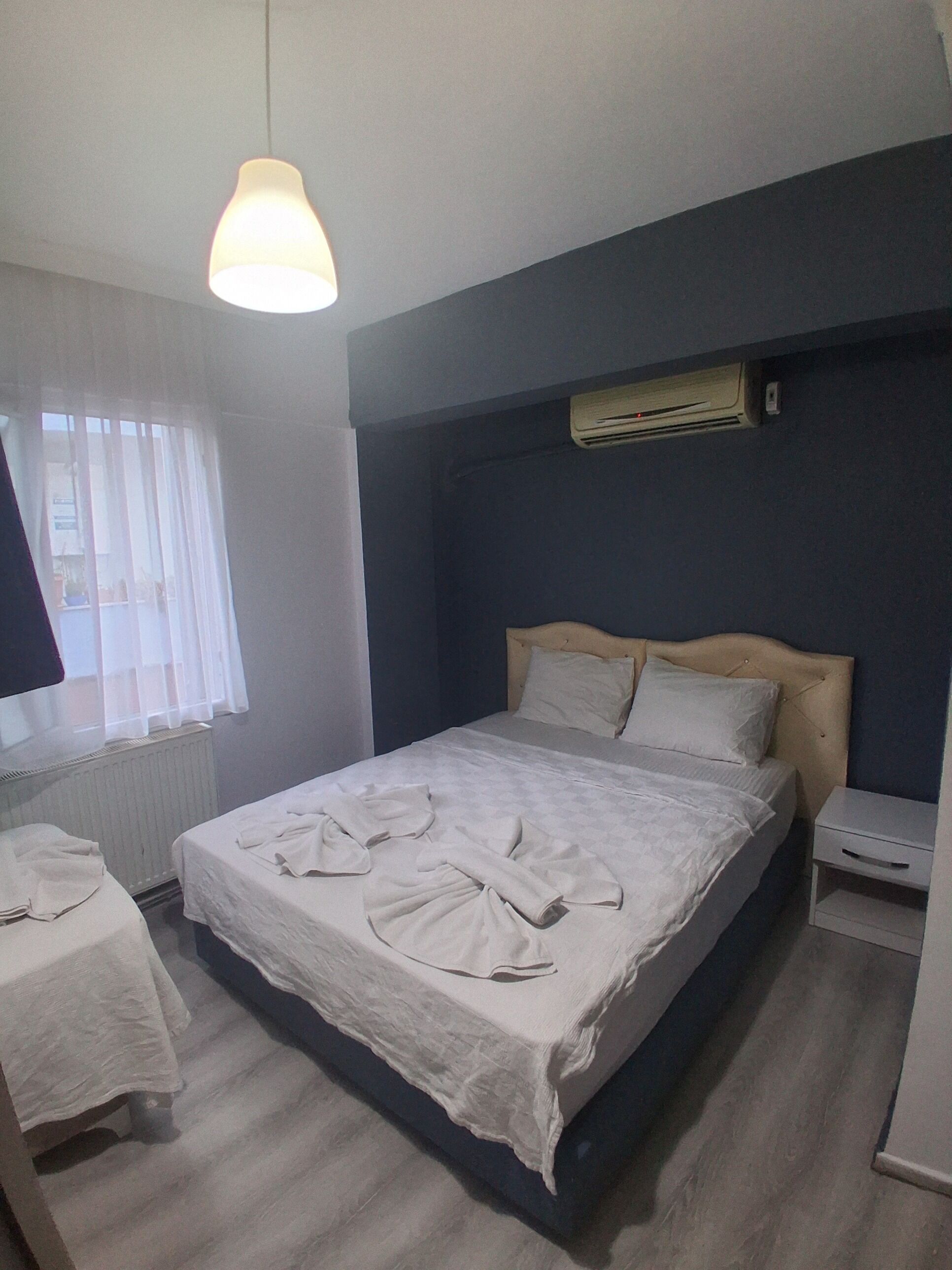 A bedroom with a bed, a bedside table, a hanging light, a window with blinds, and an air conditioning unit.
