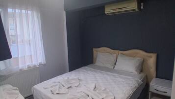 A bedroom with a bed, a bedside table, a hanging light, a window with blinds, and an air conditioning unit.