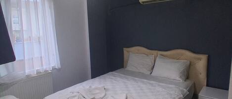 A bedroom with a bed, a bedside table, a hanging light, a window with blinds, and an air conditioning unit.