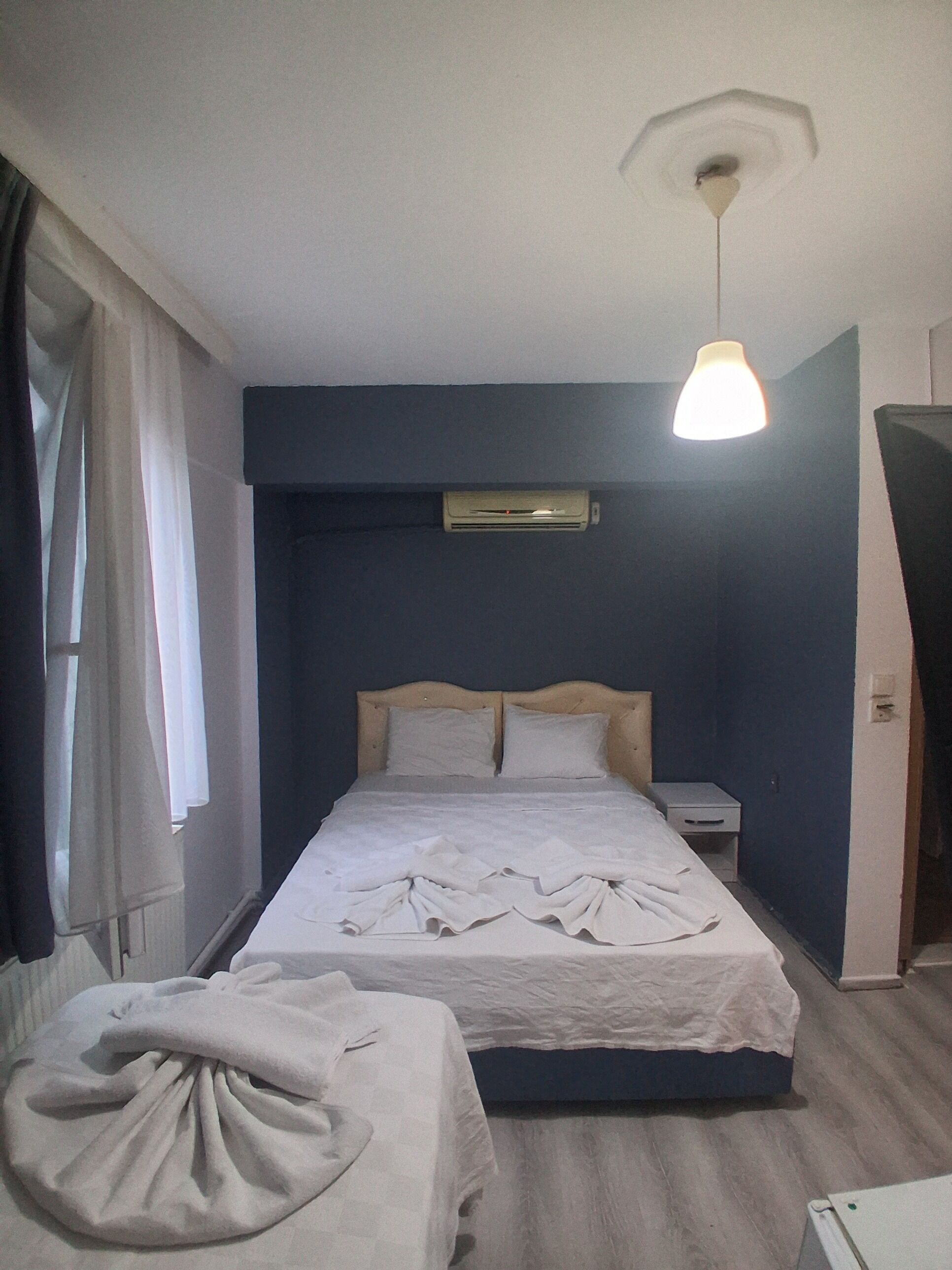 A bedroom with a bed, bedside tables, an air conditioning unit, a hanging light fixture, and a window with curtains.