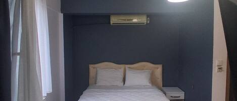 Un dormitorio con cama, mesitas de noche, equipo de aire acondicionado, lámpara colgante y ventana con cortinas.