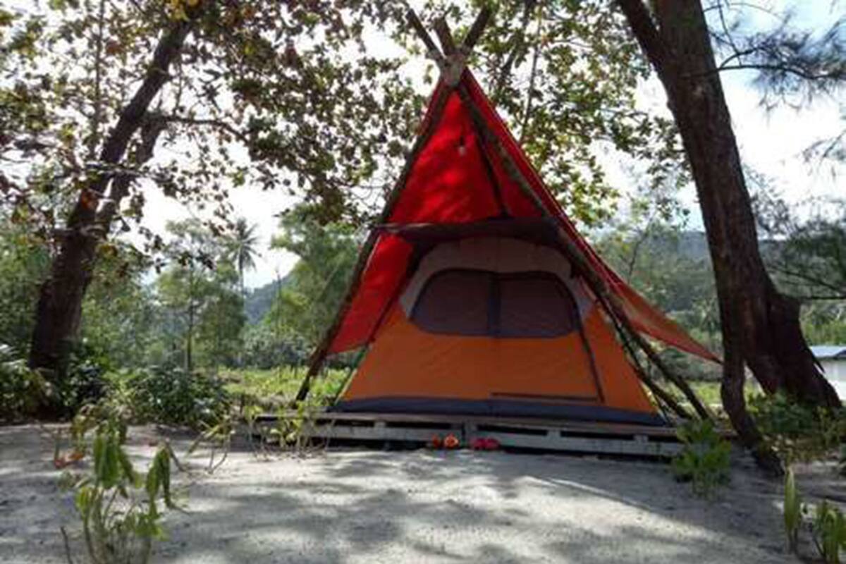 A red and orange tent set up in a forested area with trees and a gravel ground.