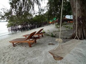 A beach with wooden lounge chairs and a hammock under a large tree.