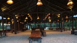 An indoor space with a high ceiling, wooden beams, and multiple hanging light fixtures. Tables and benches are arranged for seating. - Koh Rong Love Resort - Hostel (Koh Rong)