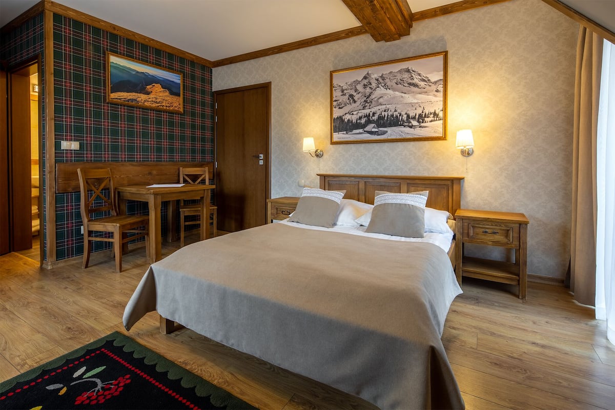 Un dormitorio con cama, cabecera de madera, mesitas de noche, un escritorio con sillas, una pared estampada con diseño escocés y dos cuadros enmarcados.