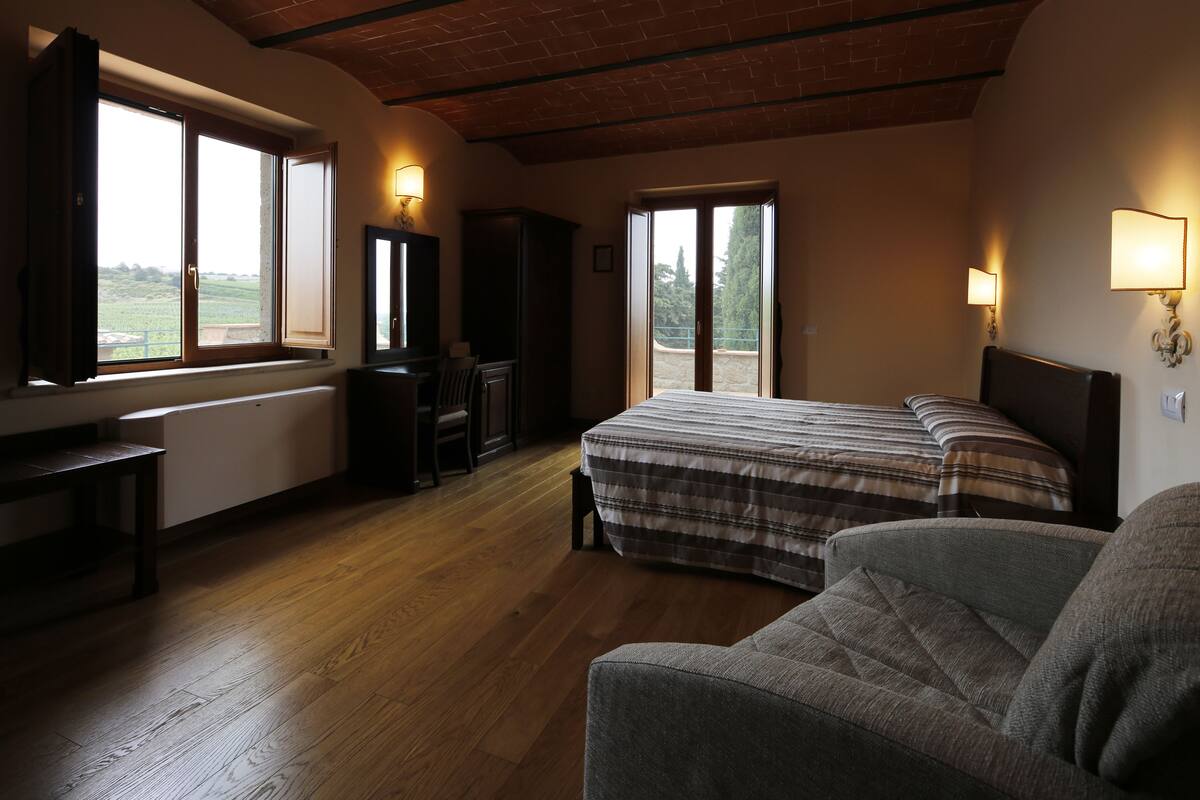 A bedroom with a bed, a chair, a desk, and a wardrobe. There are windows with views of the outdoors.