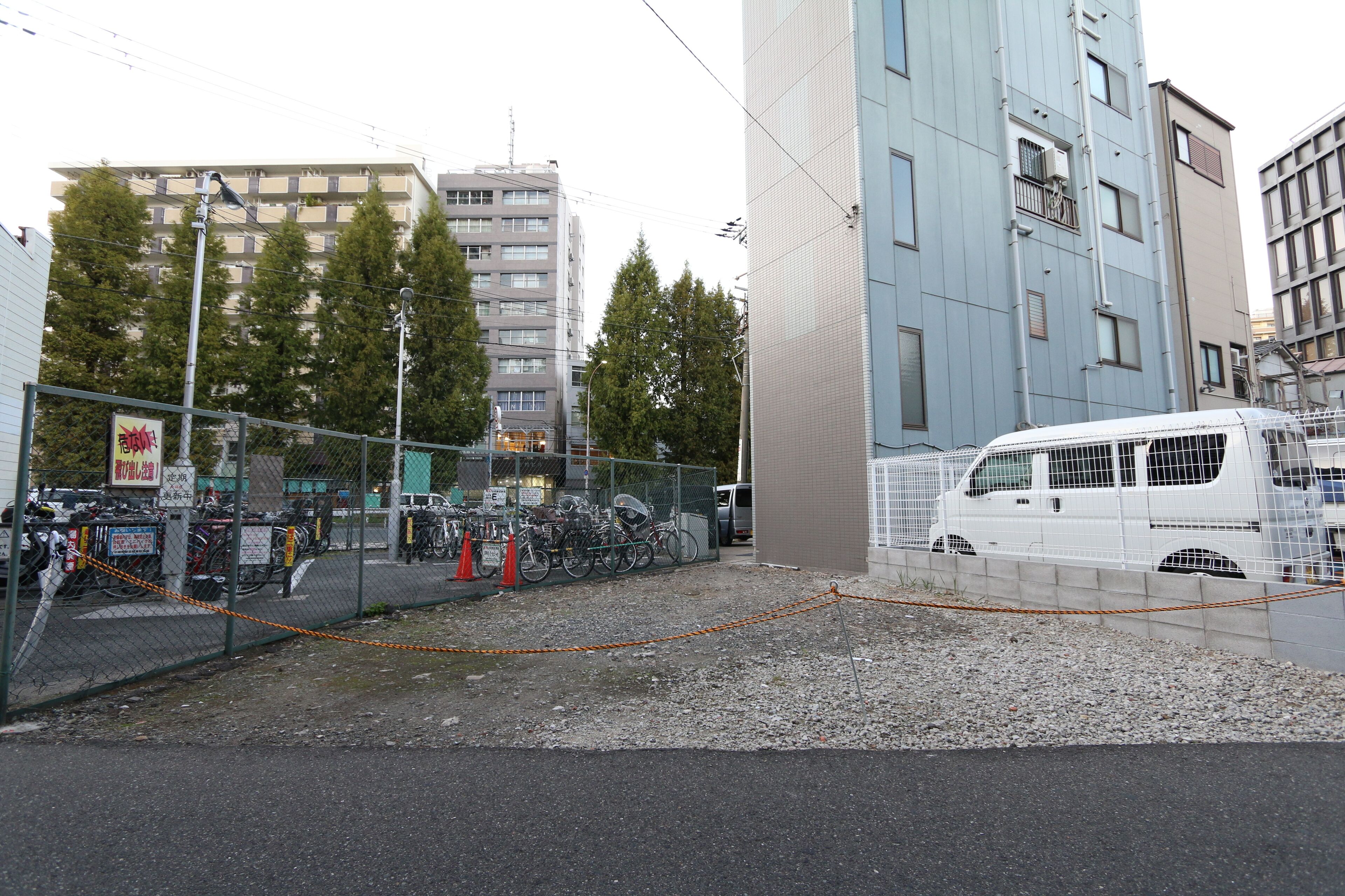 A fenced parking area with bicycles, a white van, and a building with multiple floors.