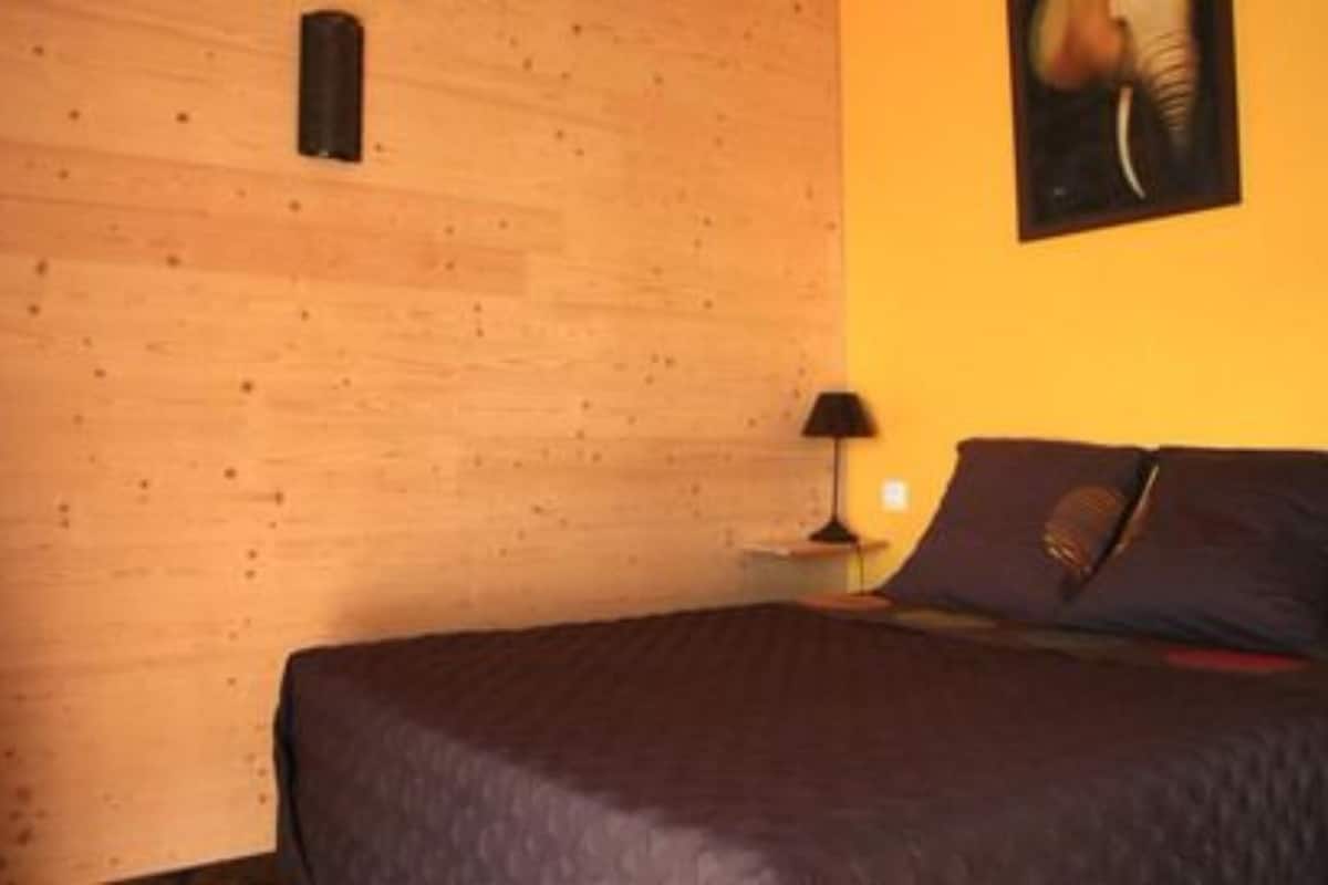 A bedroom with a wooden wall, a bed with dark bedding, a nightstand with a lamp, and a framed picture on the wall.