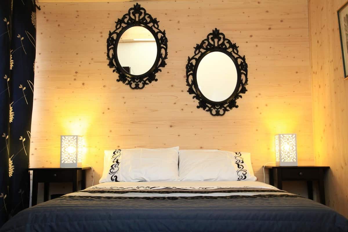 A bedroom with a wooden headboard, two ornate mirrors, white bedding, and two bedside tables with lamps.