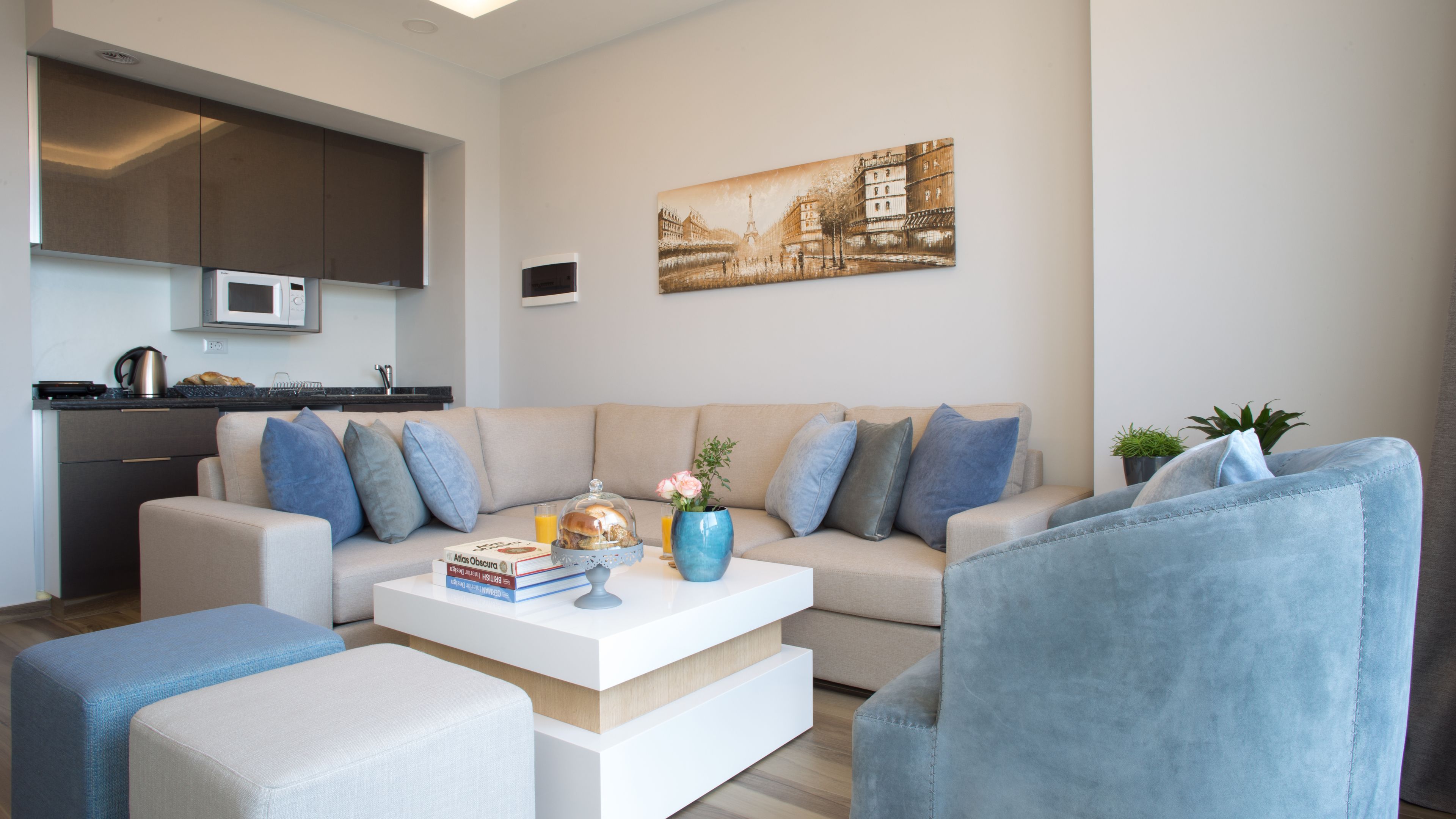 Una sala de estar moderna con sofá, mesa de centro y sillones. Hay una zona de cocina con armarios y microondas. Un cuadro de un paisaje urbano cuelga en la pared.