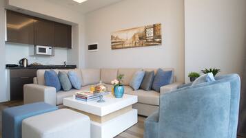 Una sala de estar moderna con sofá, mesa de centro y sillones. Hay una zona de cocina con armarios y microondas. Un cuadro de un paisaje urbano cuelga en la pared.