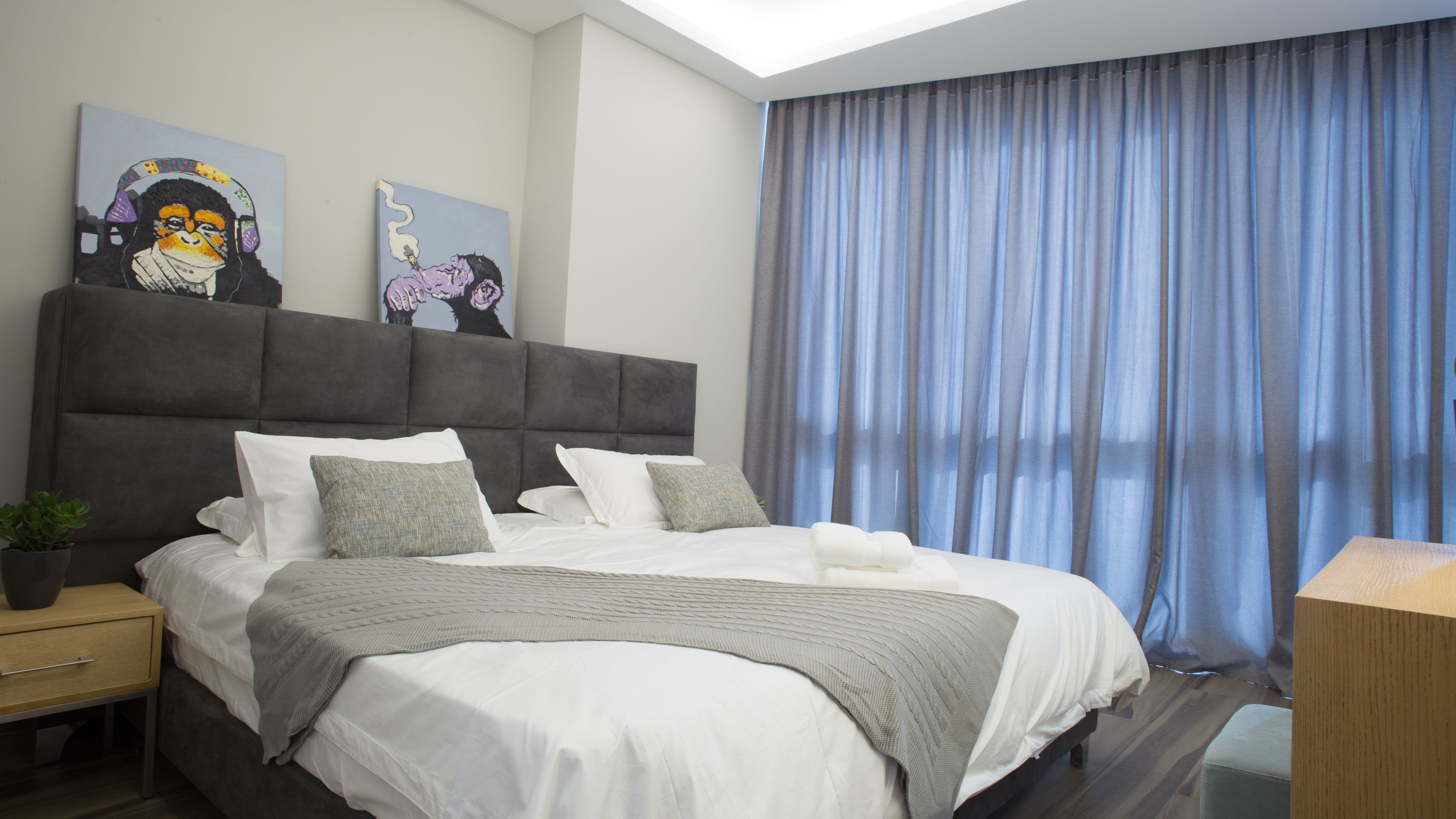 Una habitación de hotel moderna con una cama grande, una mesita de noche, un escritorio y un amplio ventanal con cortinas.