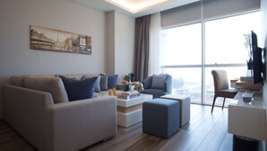 A modern living room with a sofa set, a coffee table with books and a glass of orange juice, and a view of a cityscape through large windows. - The Stay Furnished Apartments (Dbayeh)