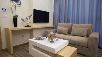A modern living room with a beige sofa, a white coffee table, a flat-screen TV mounted on the wall, and a wooden desk with a potted orchid.