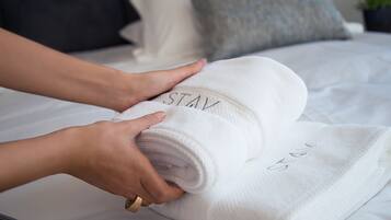 Eine Person faltet ein weißes Handtuch mit dem Schriftzug 'STAY' auf einem Bett, auf dem ein graues und ein weißes Kopfkissen liegen.