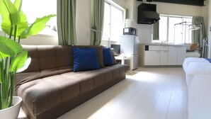 Sala de estar com um sofá marrom, uma poltrona branca e um espaço de refeições com mesa e cadeiras brancas. Há uma grande planta em vaso no lado esquerdo.