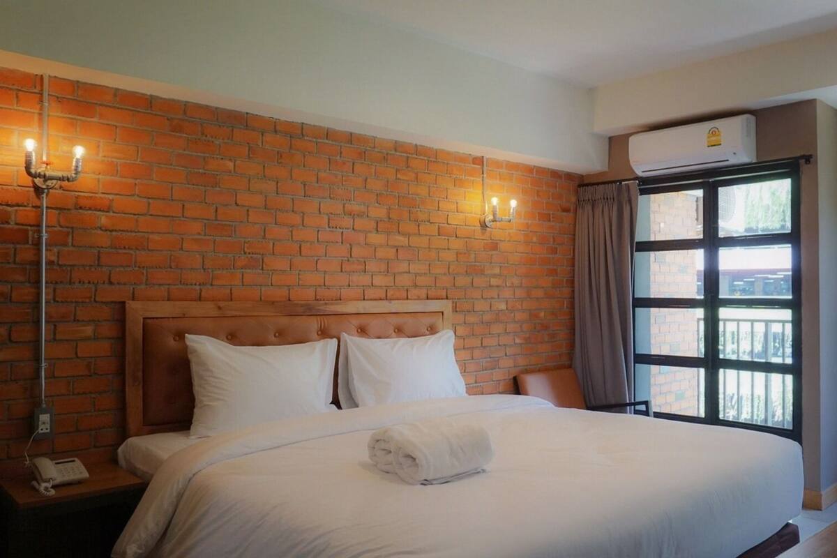 Une chambre d’hôtel avec un mur en briques, un grand lit avec du linge de lit blanc, une table de chevet avec un téléphone et une fenêtre avec des rideaux.