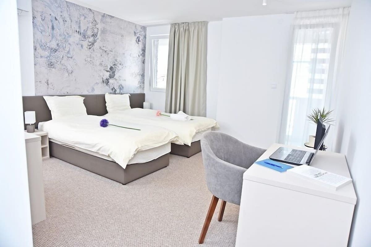 A modern hotel room with a large bed, a desk with a laptop, and a chair.