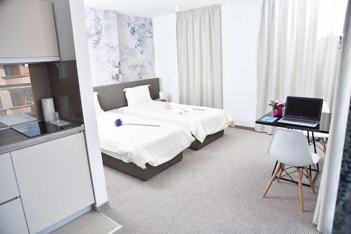 A hotel room with two beds, a desk with a laptop, a chair, and a view of the cityscape.