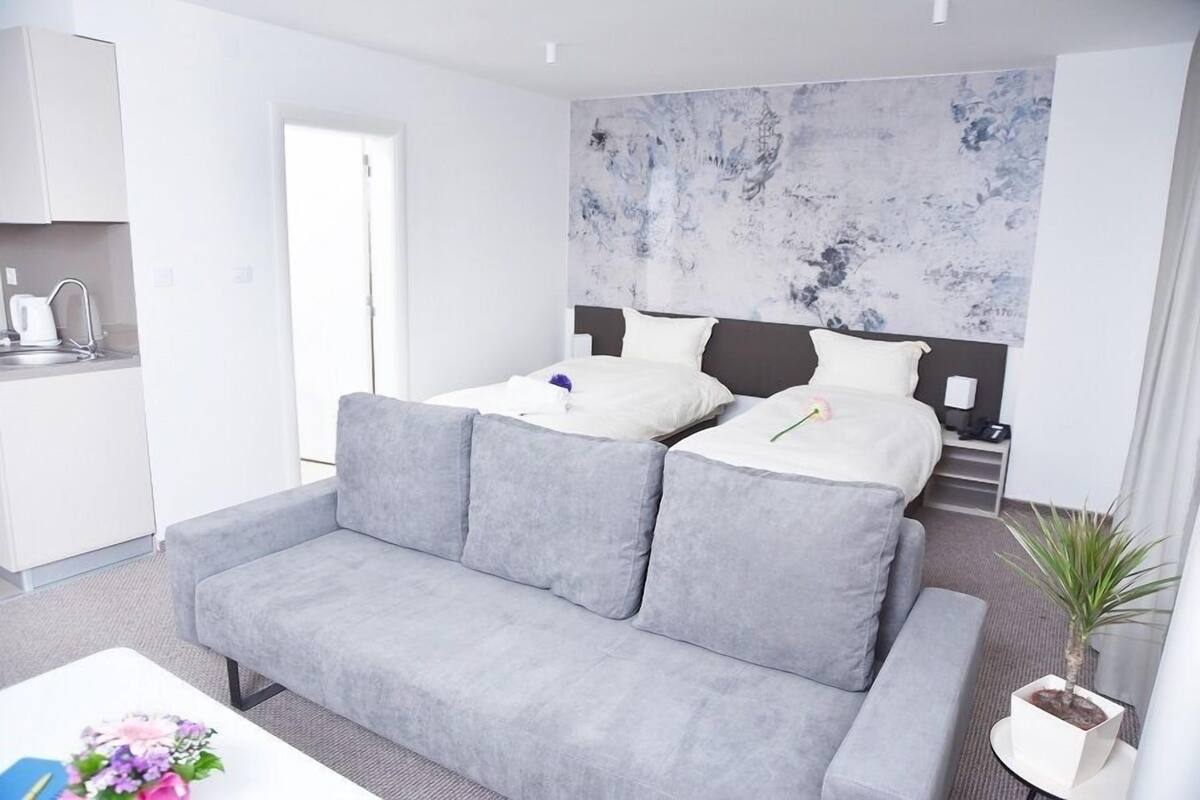 A modern hotel room with a grey sofa, a bed with white bedding, a marble-patterned wall, a small kitchen area, and a potted plant.
