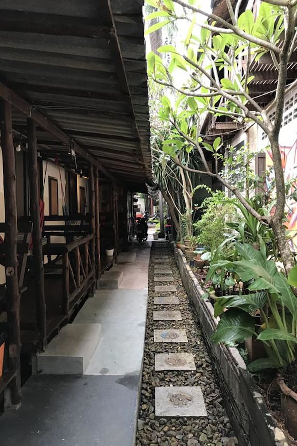 A narrow pathway flanked by wooden structures and lush greenery, leading to a building entrance.