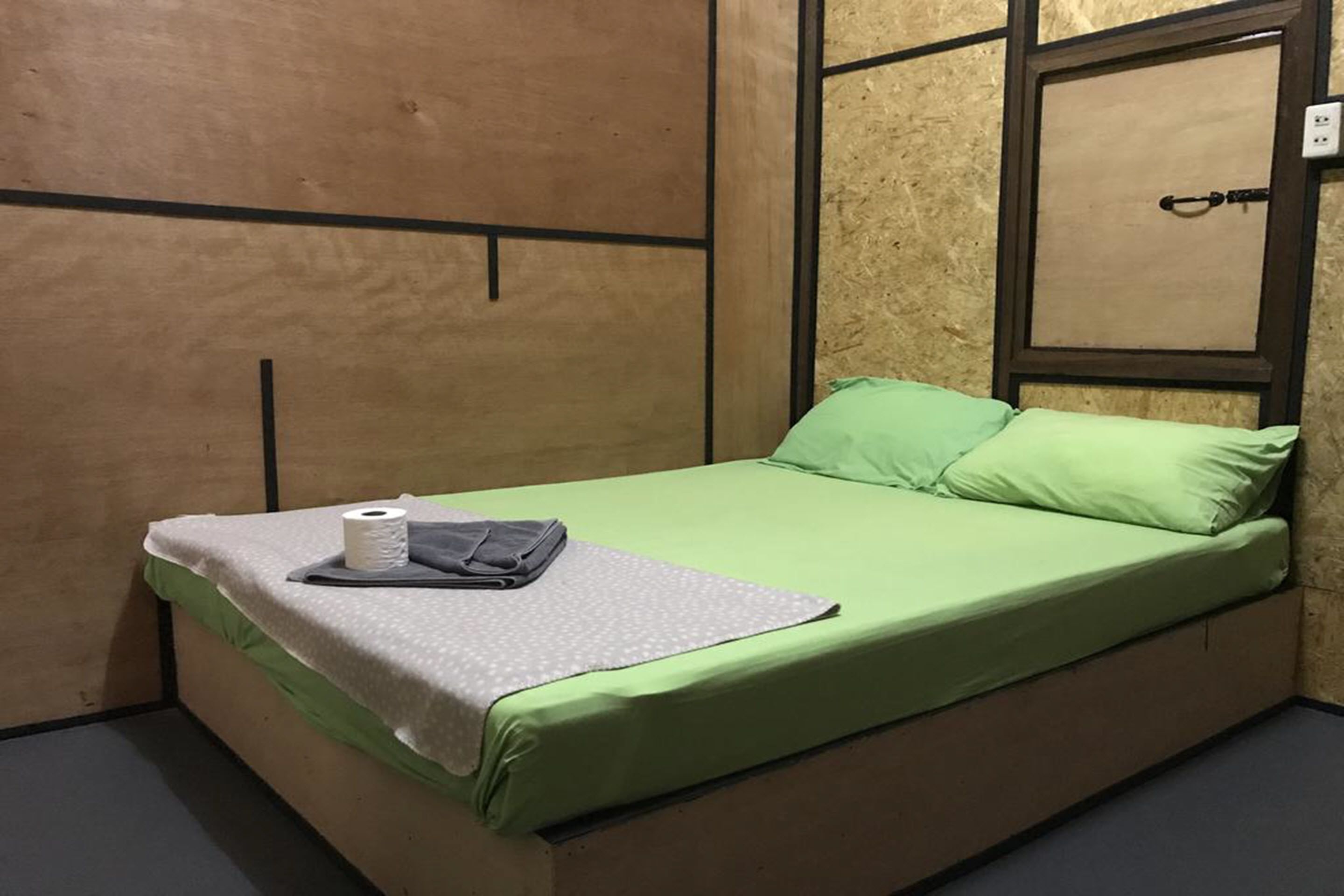 A single bed with green bedding, a pillow, a blanket, and a roll of toilet paper.