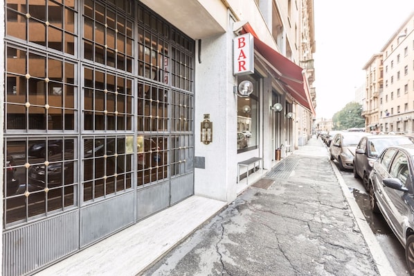 A bar with a red awning, parked cars, and a building with a grid-patterned window.