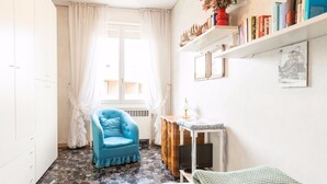 A small, well-lit room with a blue armchair, a small table with a lamp, a bed with a green blanket, and a bookshelf with various books and decorative items. - Montebello 7 (Bologna)