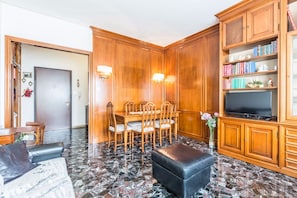 A living room with a wooden dining table, chairs, a black leather ottoman, a flat-screen TV, and a bookshelf.
