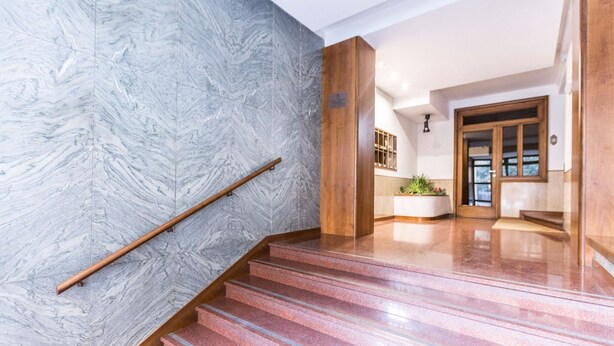 A staircase with marble walls and wooden handrails, leading to a reception area with a plant and a door. - Montebello 7 (Bologna)