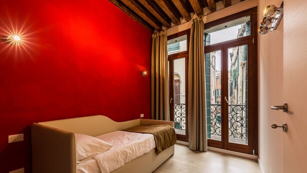 A room with a red wall, a wooden ceiling, a bed, a window with curtains, and a view of buildings outside. - Charming Palace Corte Del Teatro (Venice)