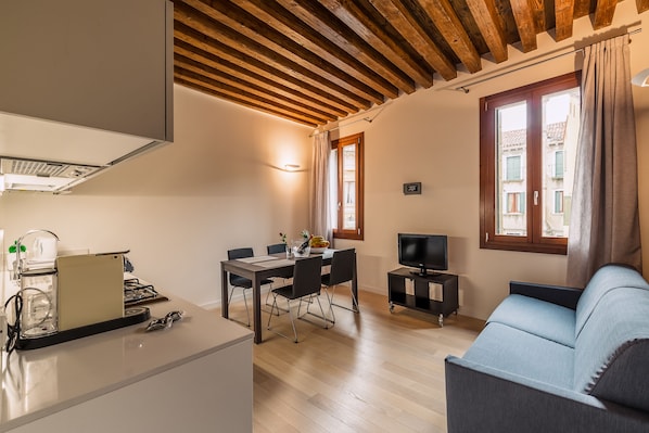 Un salon moderne avec un plafond en bois, un coin repas comprenant une table et des chaises, un canapé, un téléviseur et un coin cuisine équipé d’une cuisinière et d’une machine à café.