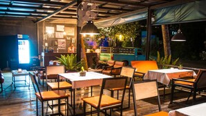 An outdoor dining area with wooden tables and orange chairs, surrounded by plants and a blue vehicle in the background.