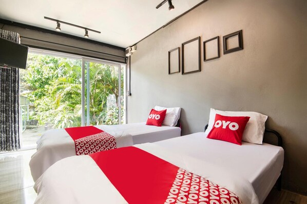 A hotel room with two beds, each with a red and white bedspread and pillows, a large window with a view of greenery, and a wall-mounted air conditioning unit.