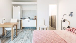 Habitación pequeña con cama, escritorio, silla, kitchenette y baño compacto.