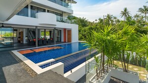 Une maison moderne à deux étages dotée d’une grande piscine, d’un espace extérieur aménagé et d’une végétation luxuriante.