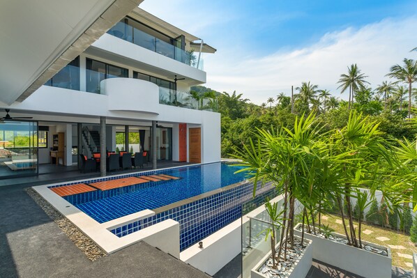 Una casa moderna de dos pisos con una gran piscina, mobiliario exterior y abundante vegetación.