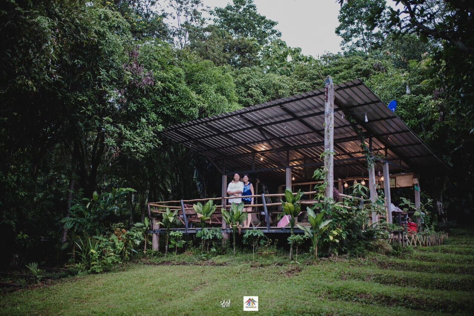 A covered outdoor area with a metal roof, surrounded by lush greenery and a small garden.