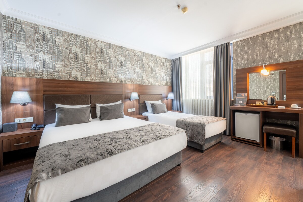 A hotel room with two beds, a desk, a mirror, and a view of the cityscape through the window.