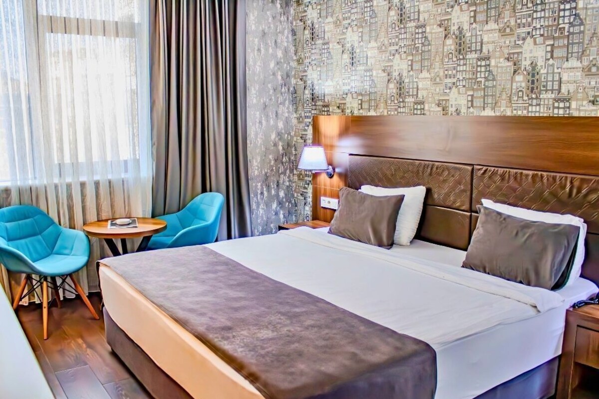 A hotel room with a large bed, two blue chairs, a small round table, and a cityscape wallpaper.