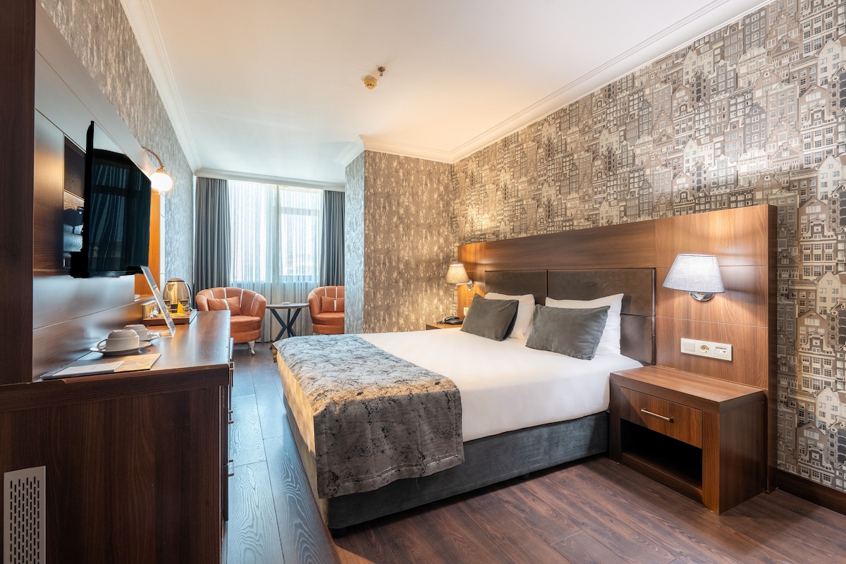 A hotel room with a large bed, bedside tables, a desk with a chair, a TV, and a patterned wallpaper.
