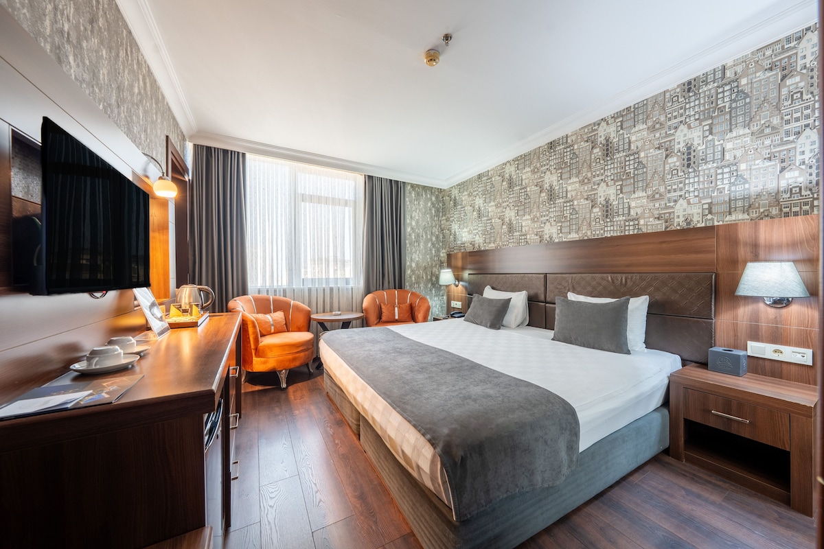 A hotel room with a large bed, two armchairs, a desk with a lamp, and a television.