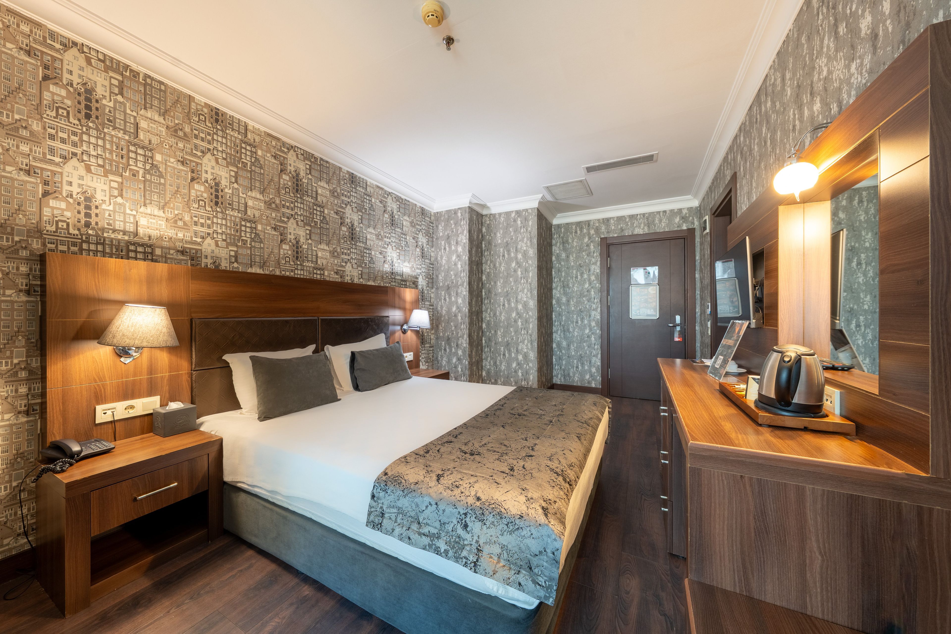 A hotel room with a large bed, bedside lamps, a wooden nightstand, a desk with a kettle, and a mirror.