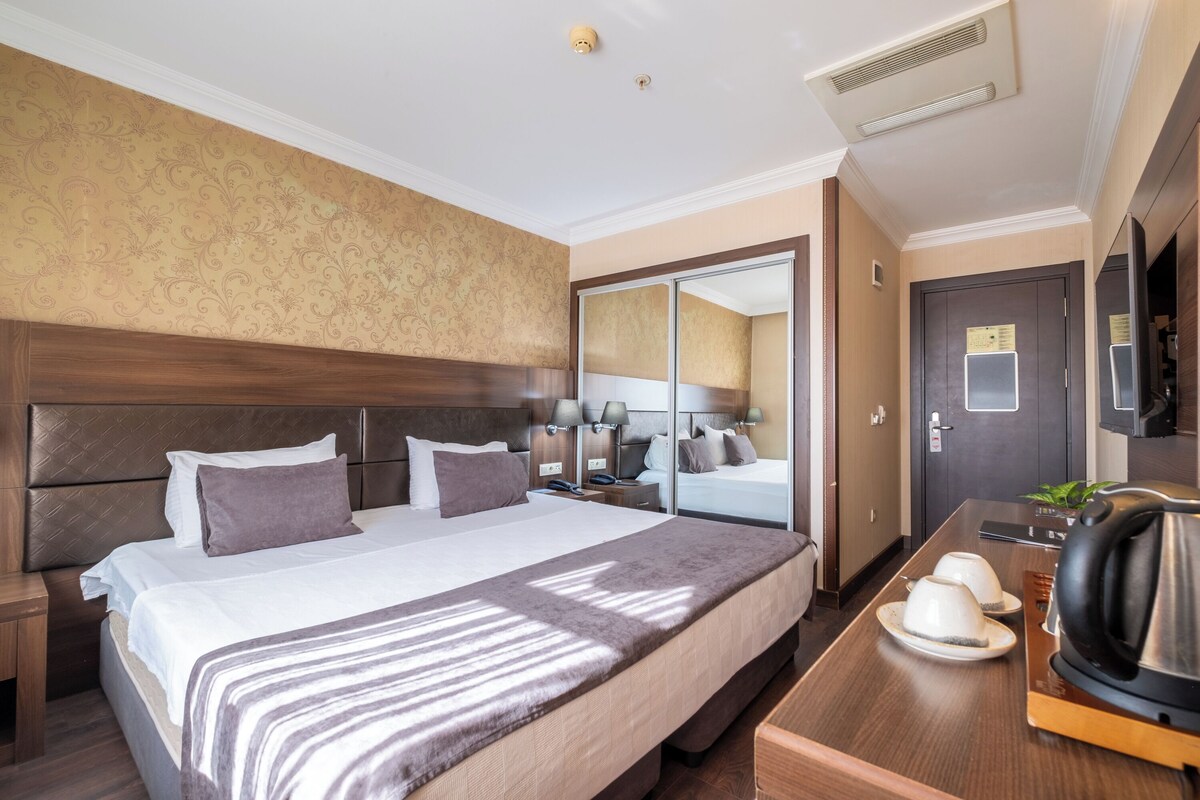 A hotel room with a large bed, a wooden headboard, a bedside table with a lamp, a mirror, a wardrobe, and a coffee maker.