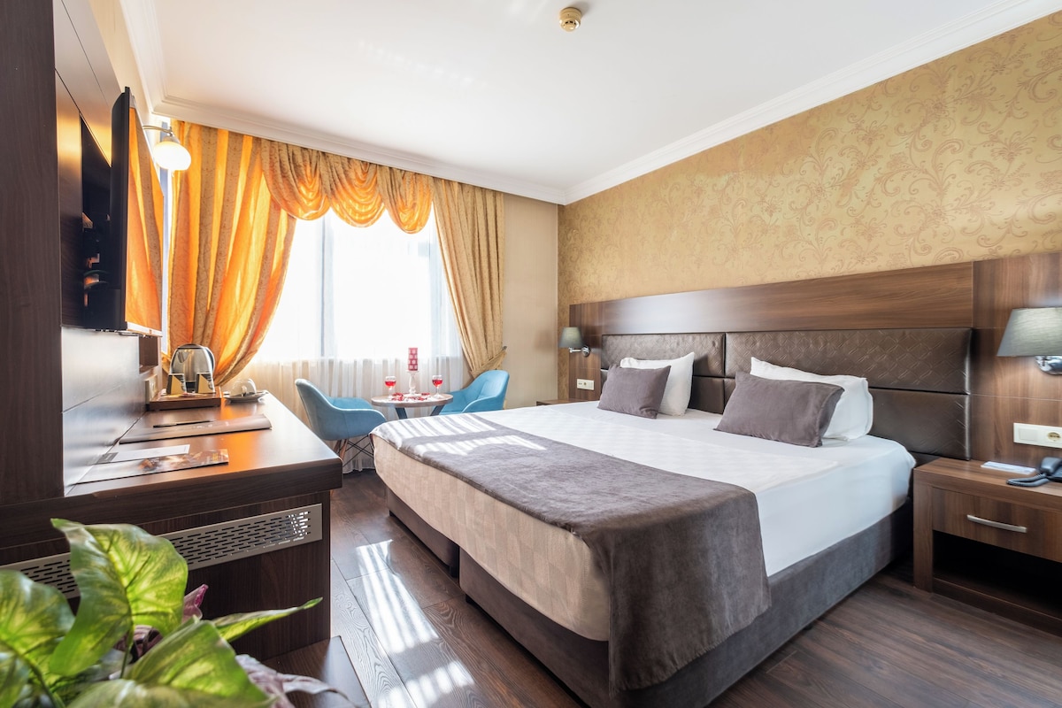 A hotel room with a large bed, a desk, a chair, and a small table with a lamp.