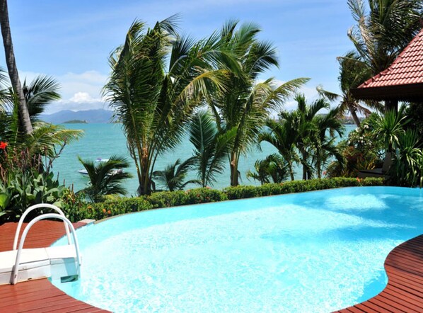 A pool with a wooden deck, surrounded by palm trees and a view of the sea.