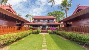 A two-story wooden house with a central balcony, surrounded by lush greenery and palm trees. - 5BR Beachfront Villa Bangrak (Koh Samui)