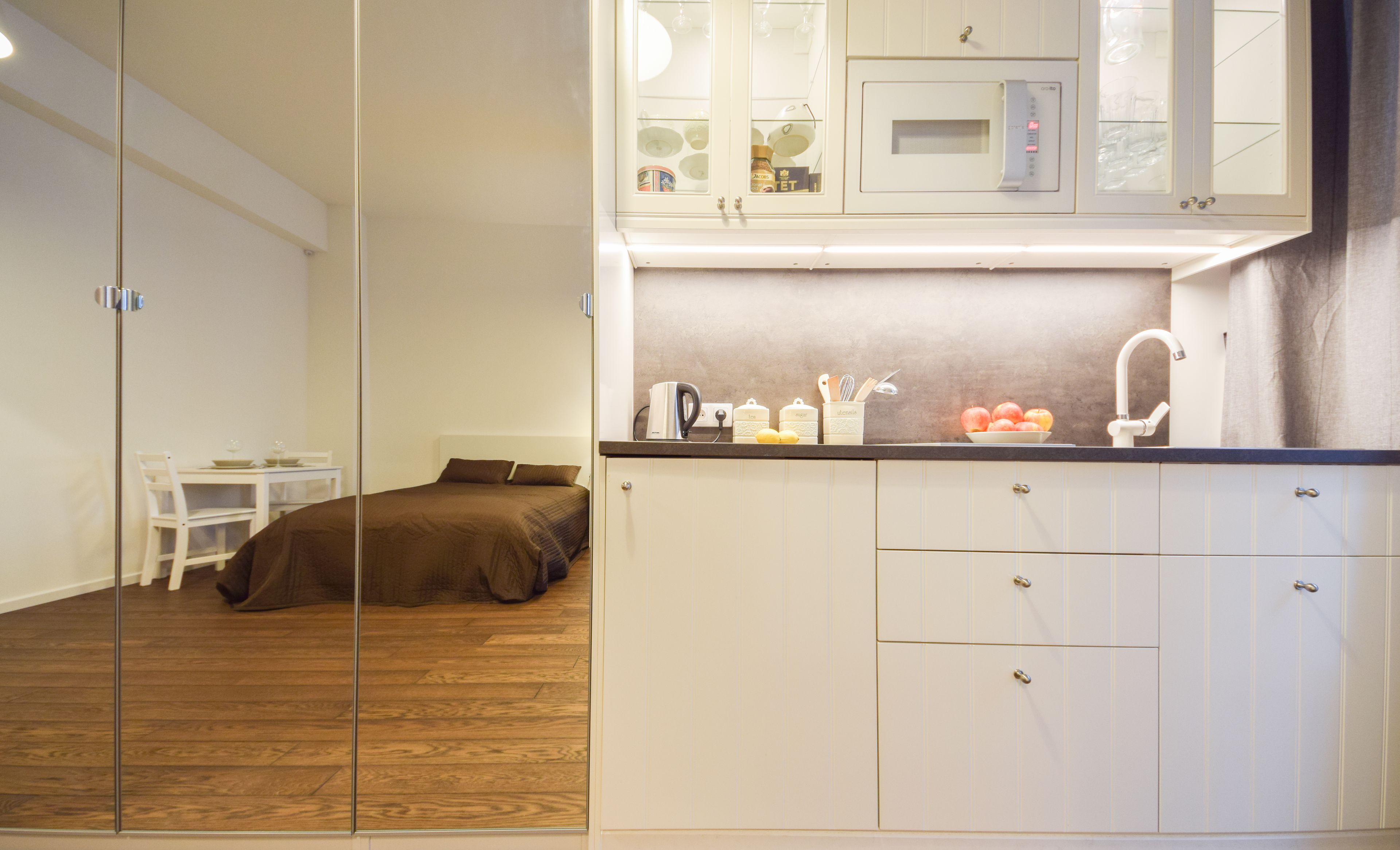 Habitación compacta con cama, una mesita pequeña y una kitchenette equipada con microondas, tetera y frutero.